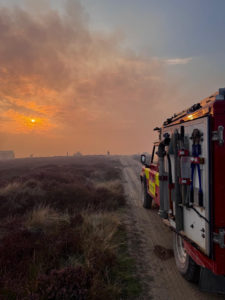 Fire and rescue vehicle- Land Rover- parked on a dirt track in open moorland during a fire response. The vehicle is on the right side of the image with equipment visible on its rear. The sky is hazy with smoke,