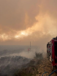 Fire and rescue vehicle positioned on a rough track at the edge of a moorland fire. Thick smoke fills the sky, creating an orange glow from the sun. In the mid-ground, a firefighter is barely visible through the smoke, with charred vegetation and patches of ash visible.